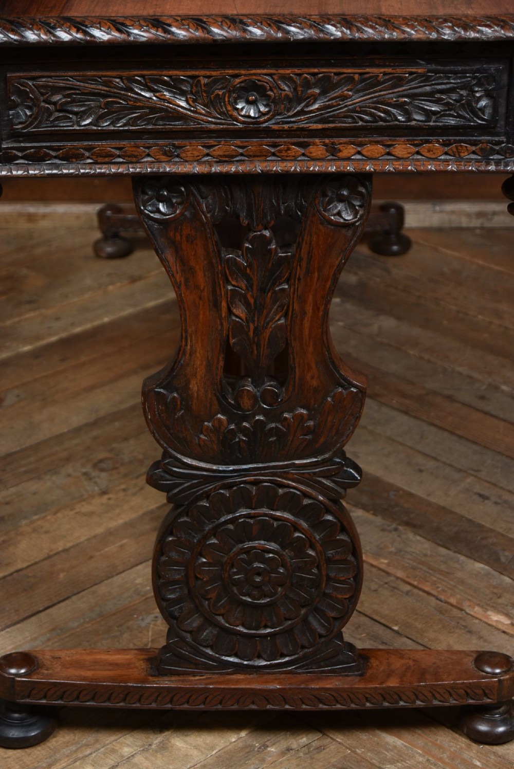 A dark brown padouk wood side table featuring a deeply carved floral gallery back, three carved drawer fronts, and trestle legs connected by a turned stretcher. View XI