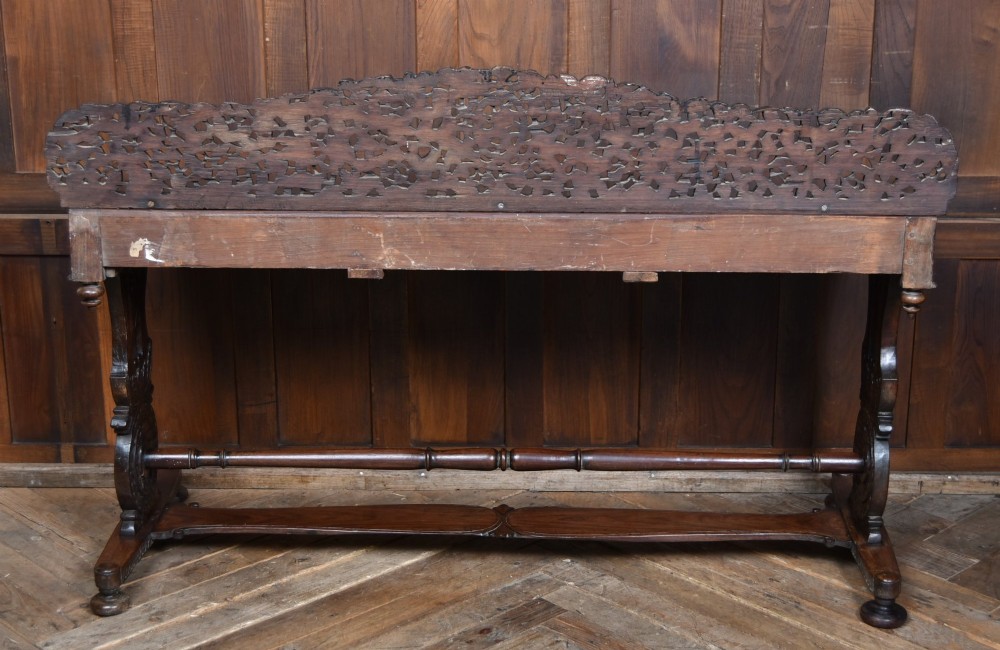 A dark brown padouk wood side table featuring a deeply carved floral gallery back, three carved drawer fronts, and trestle legs connected by a turned stretcher. View XII