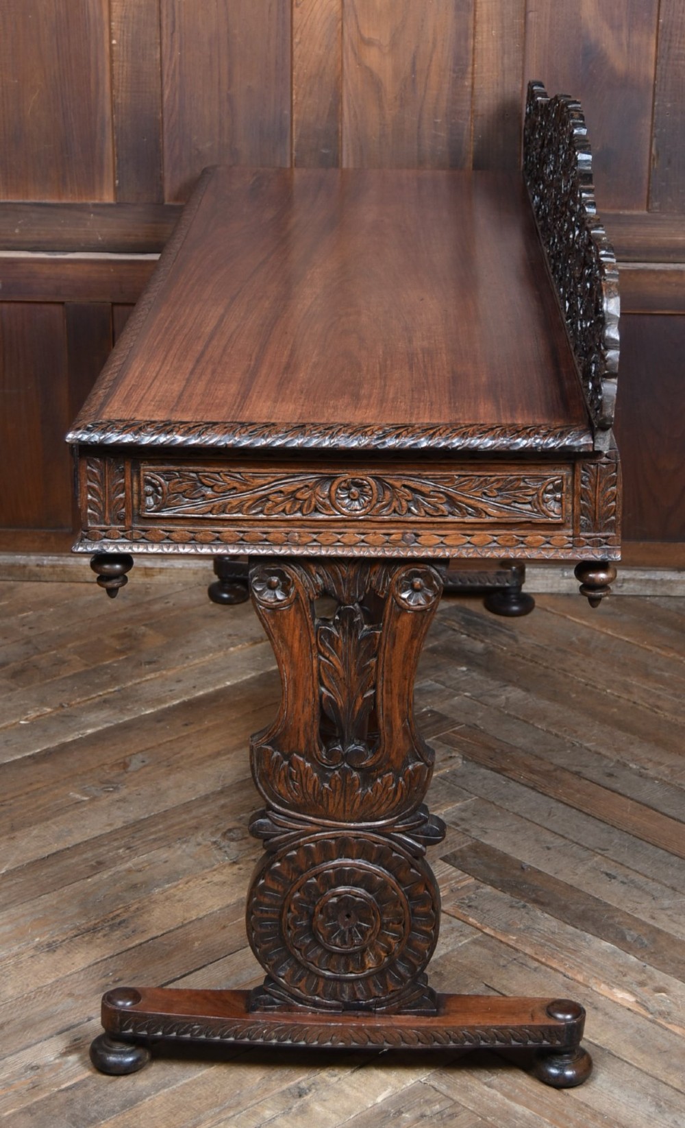 A dark brown padouk wood side table featuring a deeply carved floral gallery back, three carved drawer fronts, and trestle legs connected by a turned stretcher. View XIII