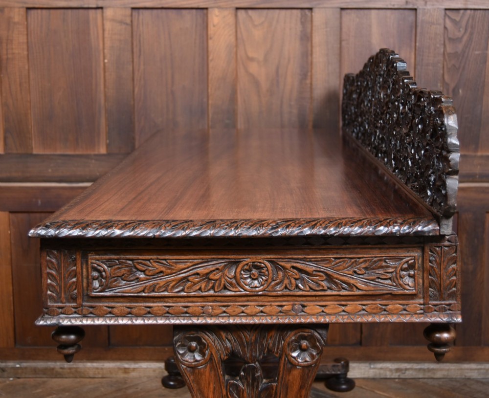 A dark brown padouk wood side table featuring a deeply carved floral gallery back, three carved drawer fronts, and trestle legs connected by a turned stretcher. View XIV