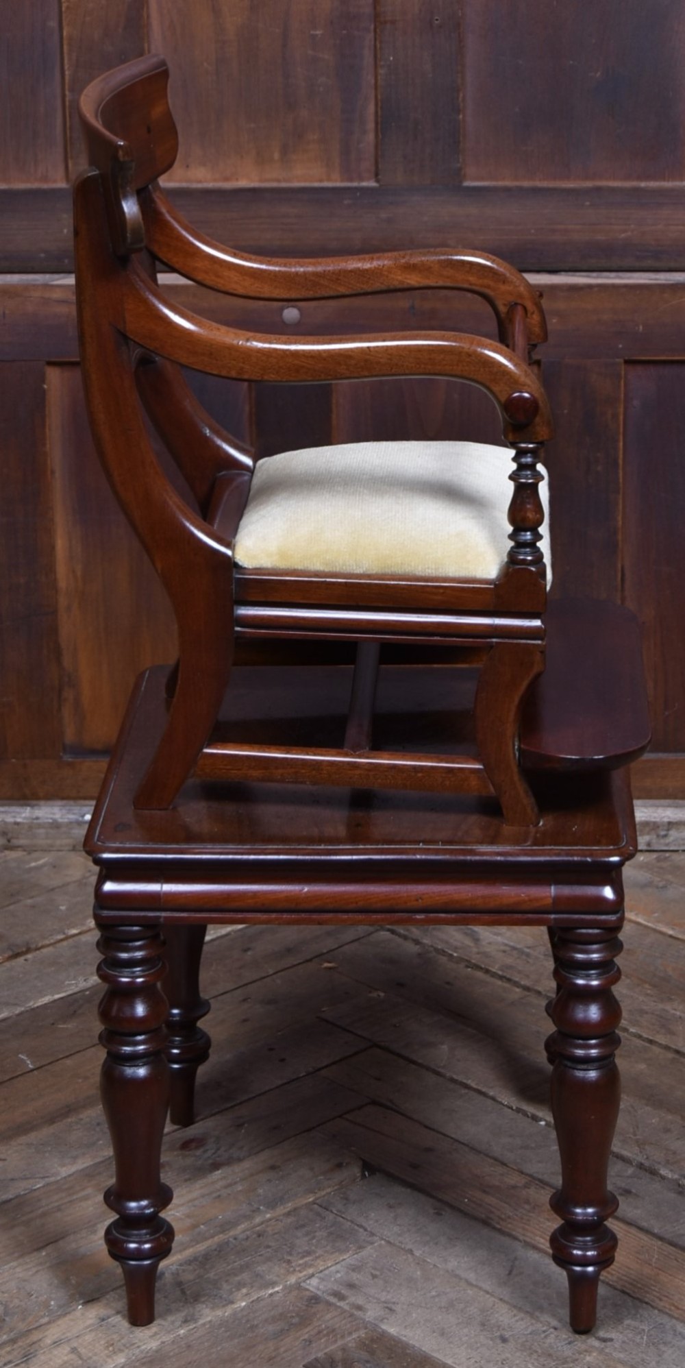A Victorian metamorphic mahogany childs high chair with turned legs, curved armrests, a carved backrest crest, and cream fabric upholstery. View V