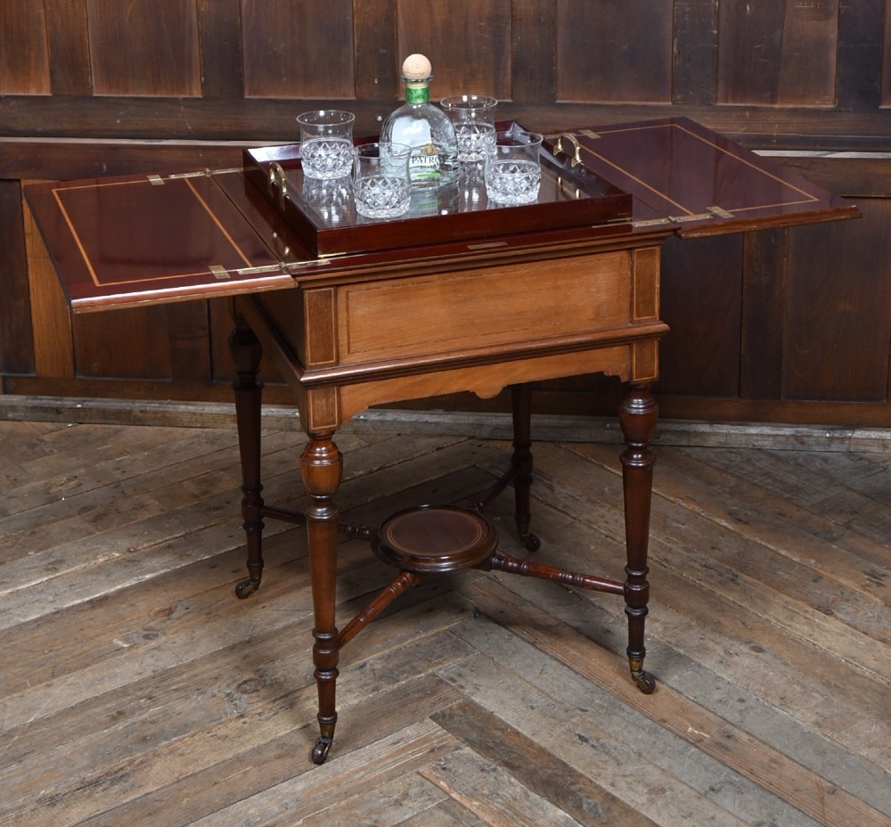 An Edwardian mahogany drinks cabinet table featuring hinged folding top leaves, a rising central tray mechanism, satinwood inlay, and turned legs on brass castors.