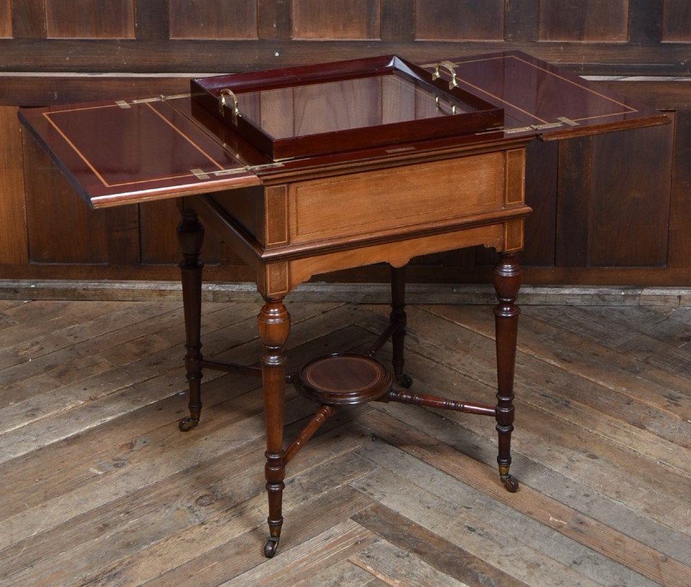 An Edwardian mahogany drinks cabinet table featuring hinged folding top leaves, a rising central tray mechanism, satinwood inlay, and turned legs on brass castors. View II