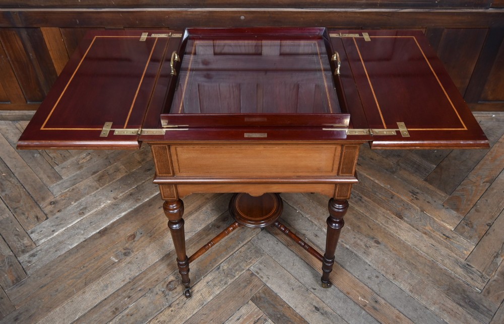 An Edwardian mahogany drinks cabinet table featuring hinged folding top leaves, a rising central tray mechanism, satinwood inlay, and turned legs on brass castors. View IX