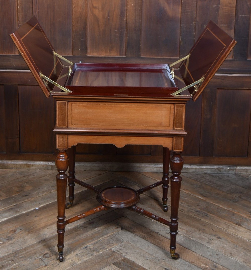 An Edwardian mahogany drinks cabinet table featuring hinged folding top leaves, a rising central tray mechanism, satinwood inlay, and turned legs on brass castors. View X