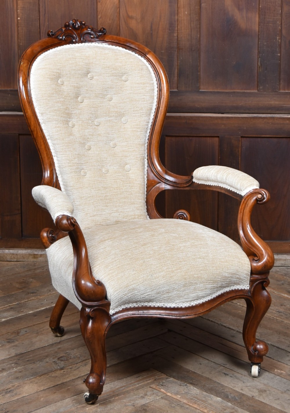 A Victorian walnut spoon-back armchair upholstered in neutral fabric with button tufting, carved scroll arms, cabriole legs, and castors.