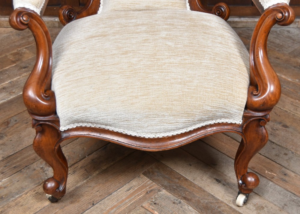 A Victorian walnut spoon-back armchair upholstered in neutral fabric with button tufting, carved scroll arms, cabriole legs, and castors. View VI