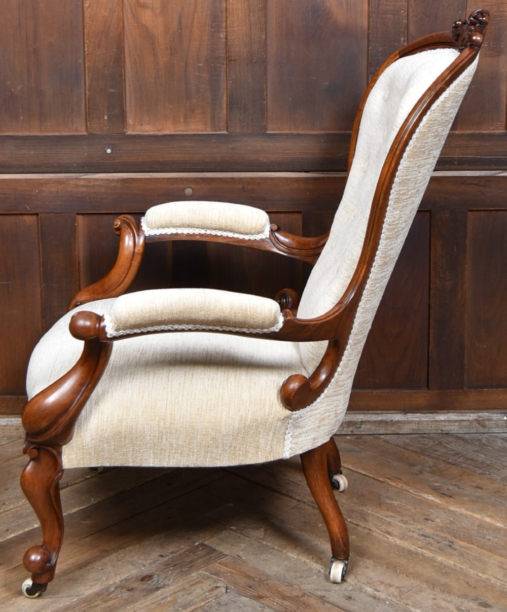 A Victorian walnut spoon-back armchair upholstered in neutral fabric with button tufting, carved scroll arms, cabriole legs, and castors. View X