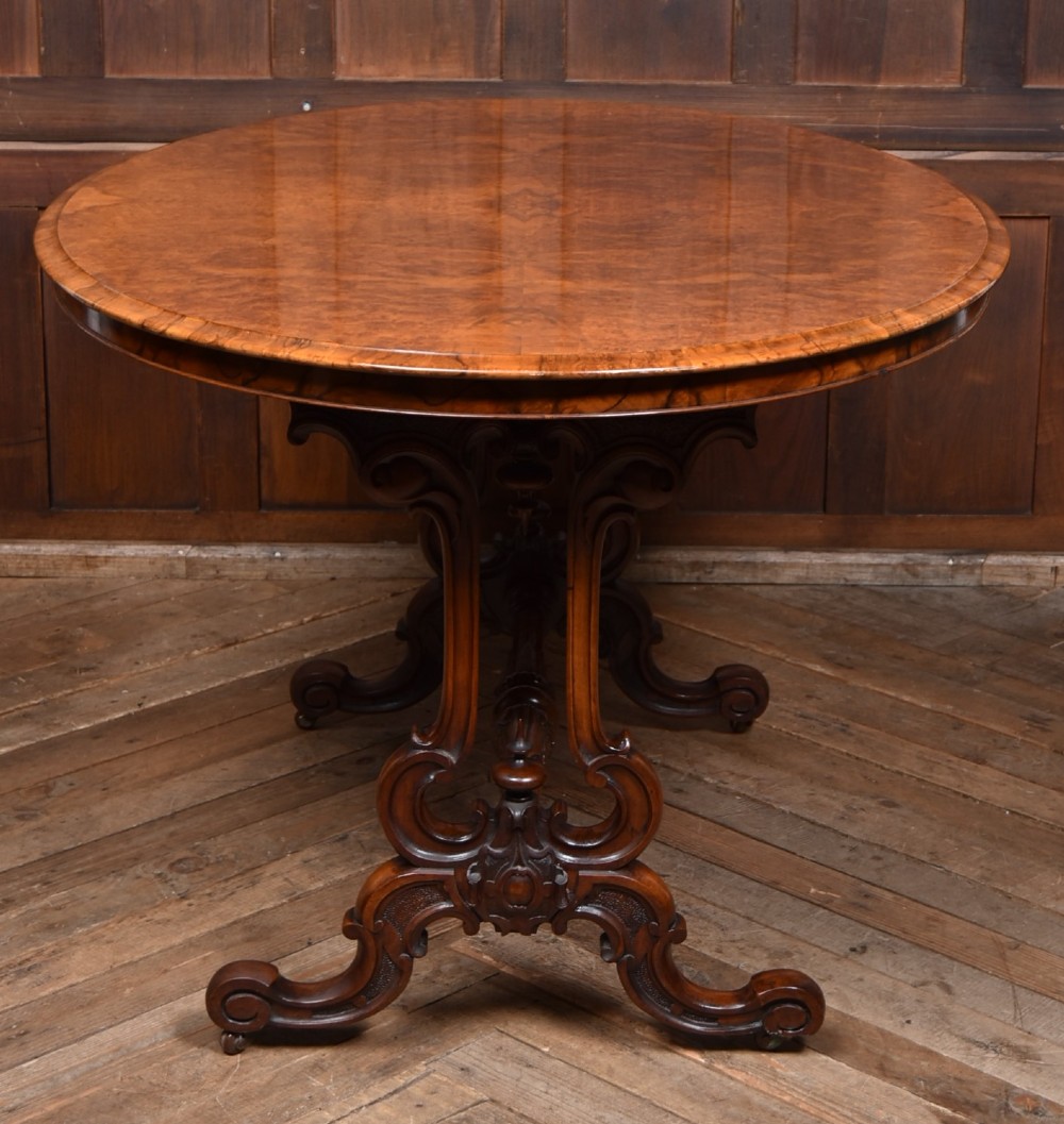 A Victorian oval centre table constructed from burr walnut, featuring carved twin pedestal legs connected by a central turned stretcher. View X