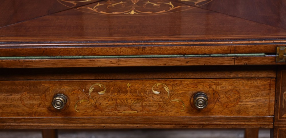 An Edwardian mahogany envelope card table featuring satinwood marquetry inlay, a single drawer, and a curved cross-stretcher with brass castors. View II