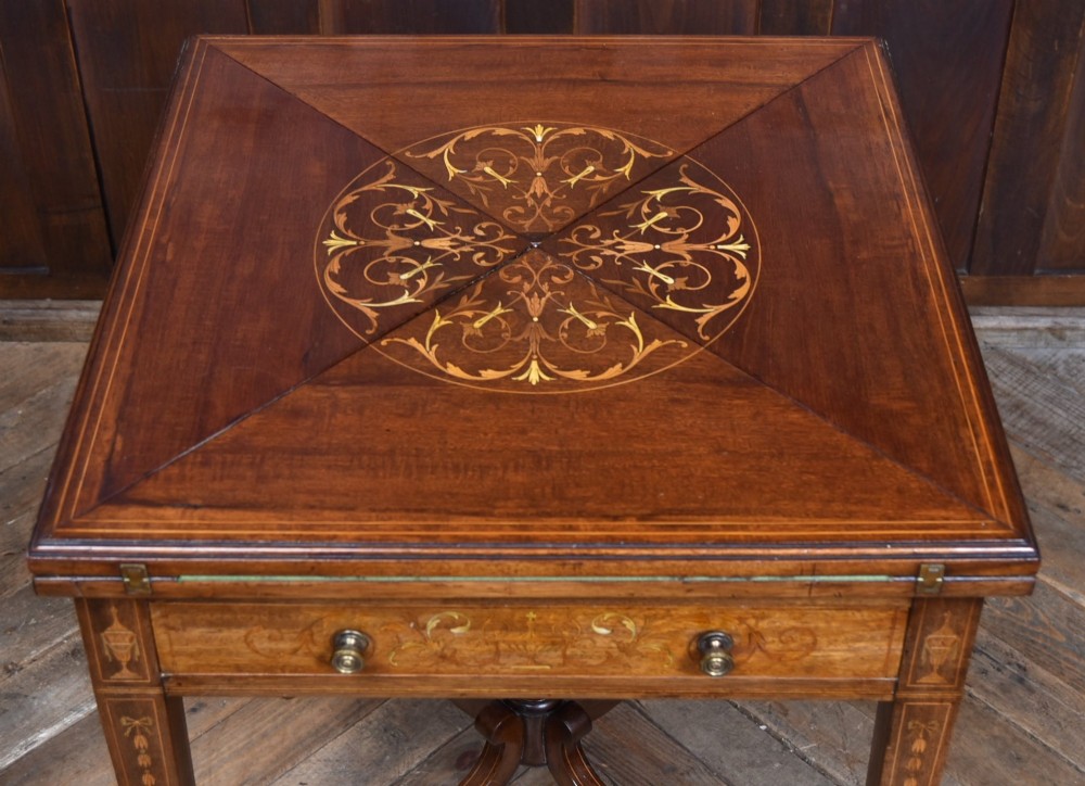 An Edwardian mahogany envelope card table featuring satinwood marquetry inlay, a single drawer, and a curved cross-stretcher with brass castors. View IV