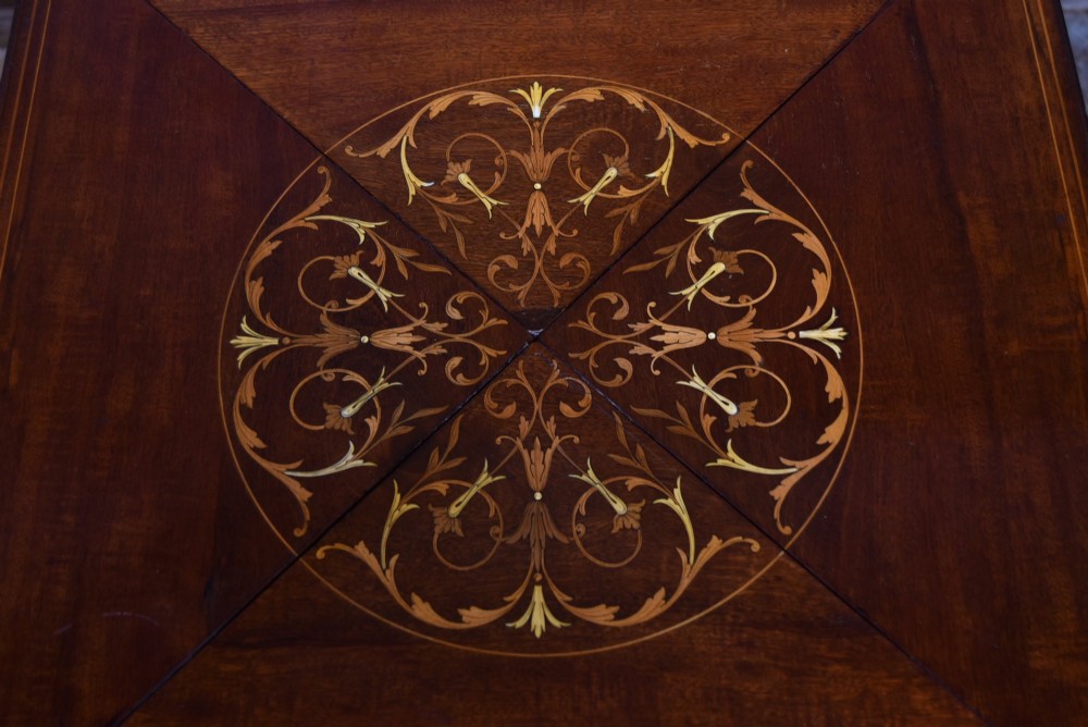 An Edwardian mahogany envelope card table featuring satinwood marquetry inlay, a single drawer, and a curved cross-stretcher with brass castors. View V