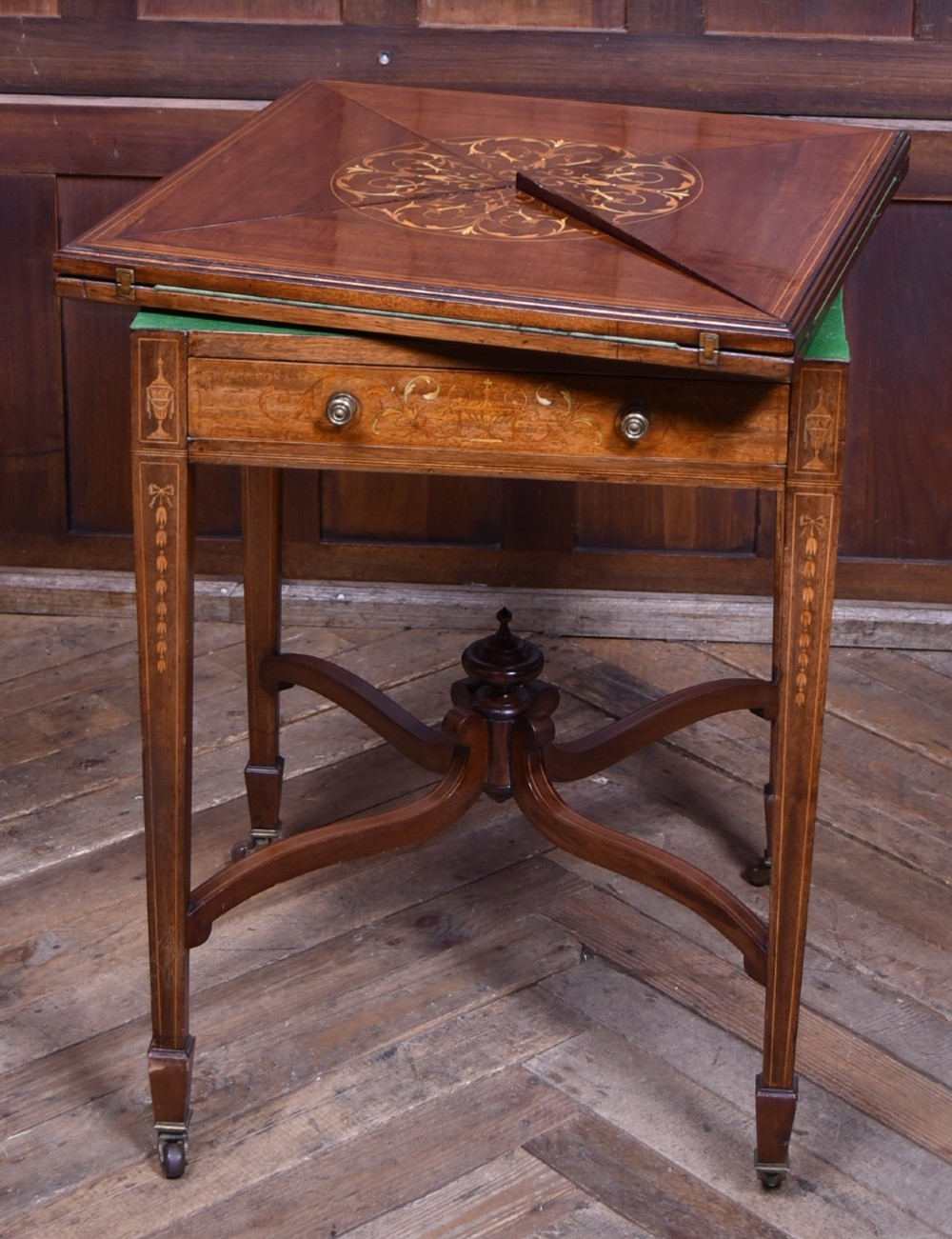 An Edwardian mahogany envelope card table featuring satinwood marquetry inlay, a single drawer, and a curved cross-stretcher with brass castors. View VI