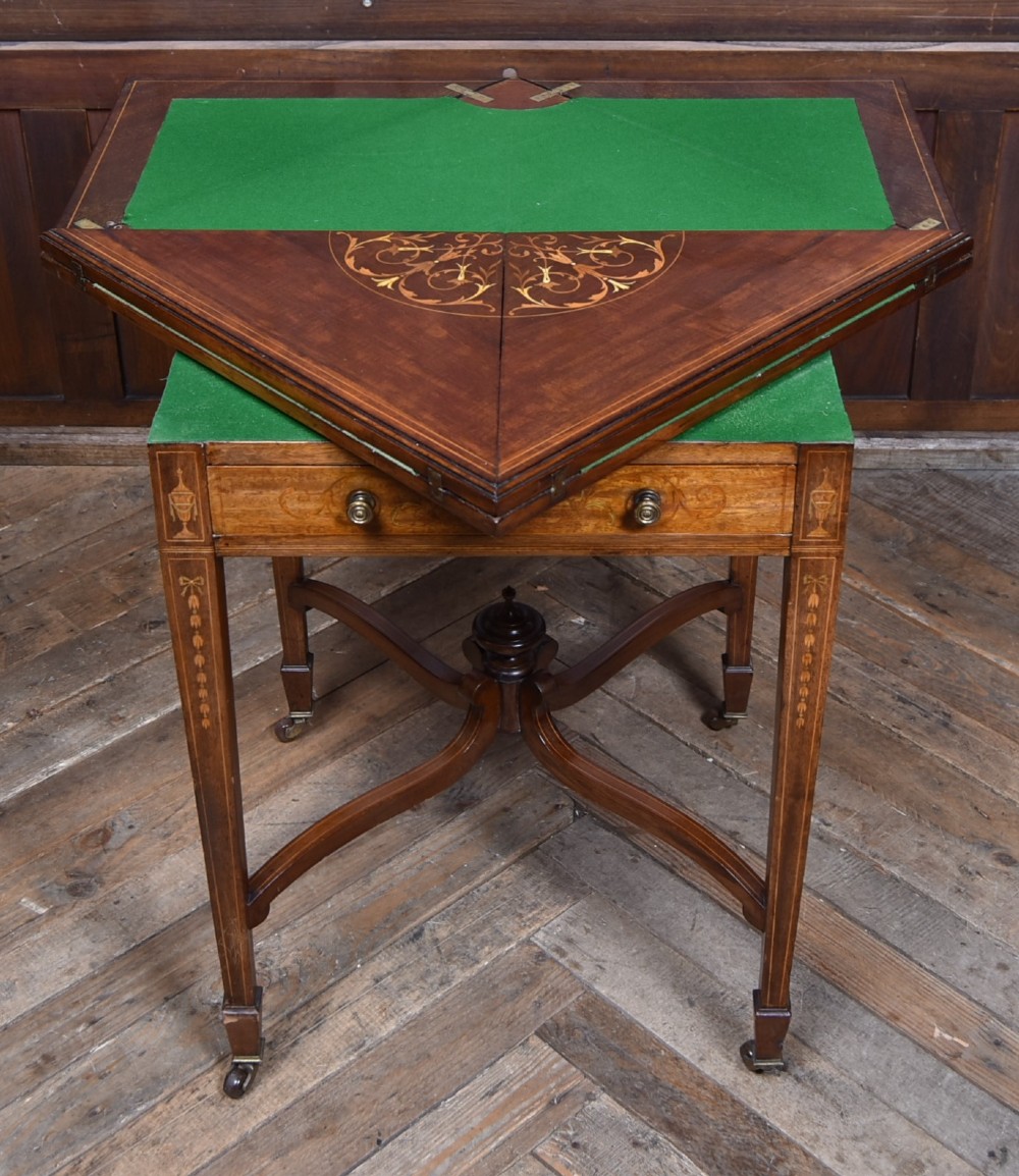 An Edwardian mahogany envelope card table featuring satinwood marquetry inlay, a single drawer, and a curved cross-stretcher with brass castors. View IX