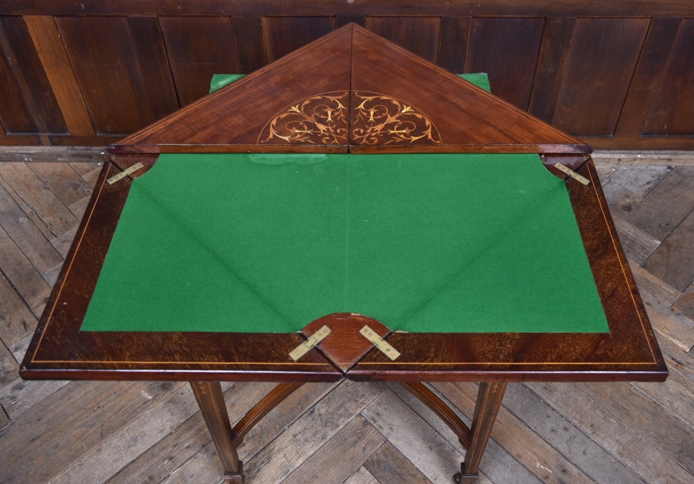 An Edwardian mahogany envelope card table featuring satinwood marquetry inlay, a single drawer, and a curved cross-stretcher with brass castors. View X