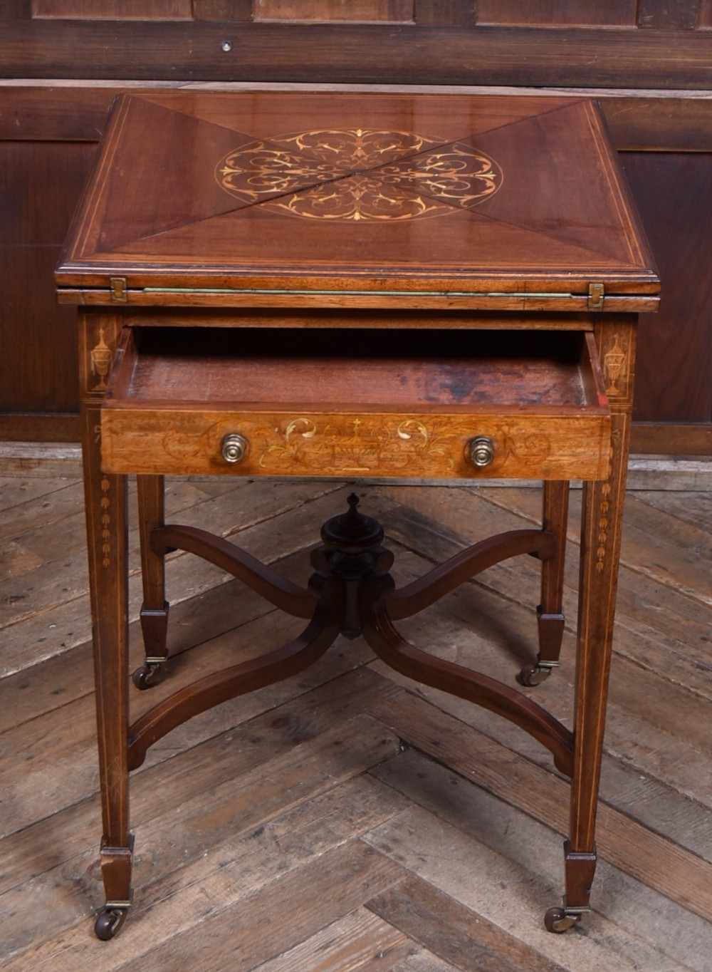 An Edwardian mahogany envelope card table featuring satinwood marquetry inlay, a single drawer, and a curved cross-stretcher with brass castors. View XI