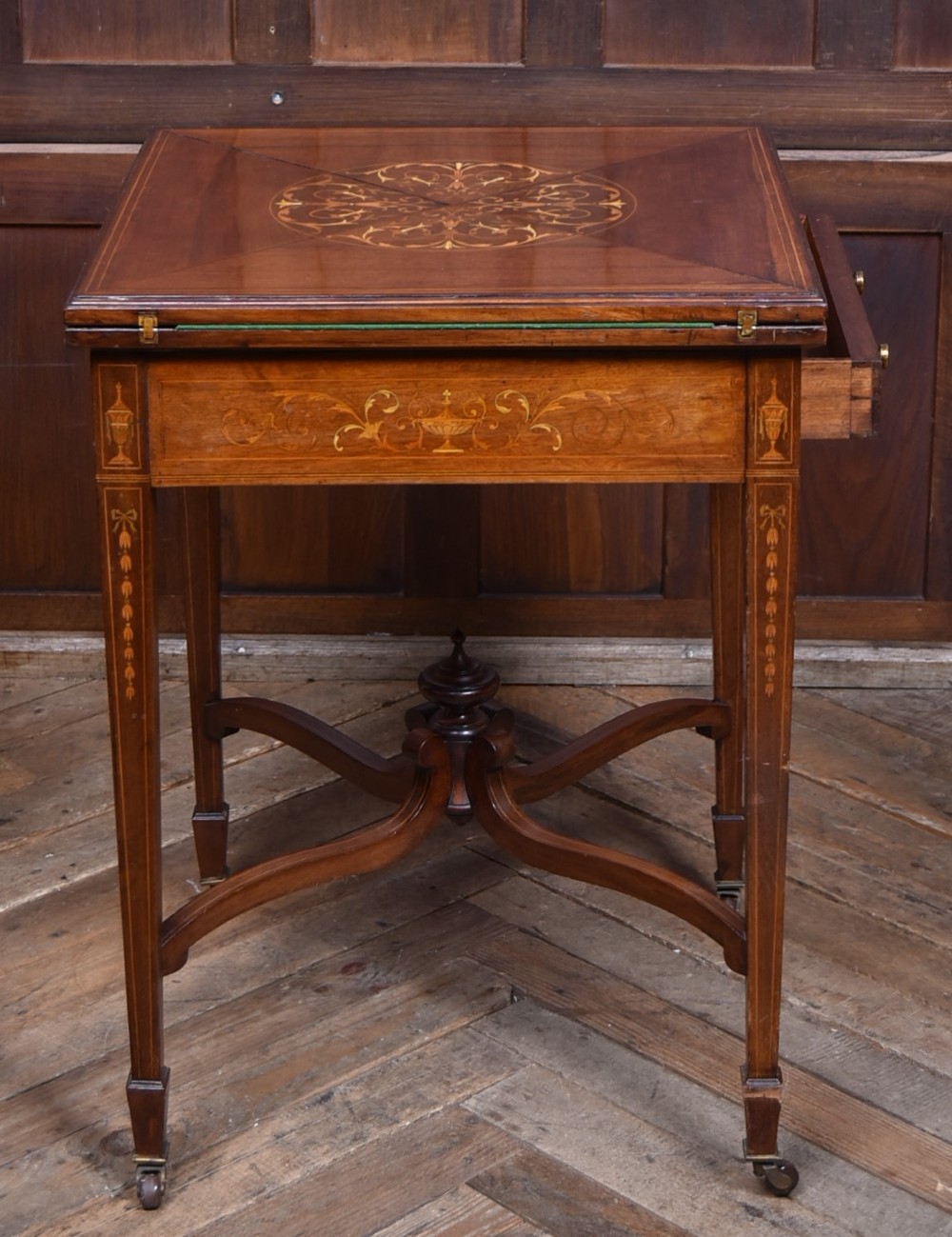 An Edwardian mahogany envelope card table featuring satinwood marquetry inlay, a single drawer, and a curved cross-stretcher with brass castors. View XII