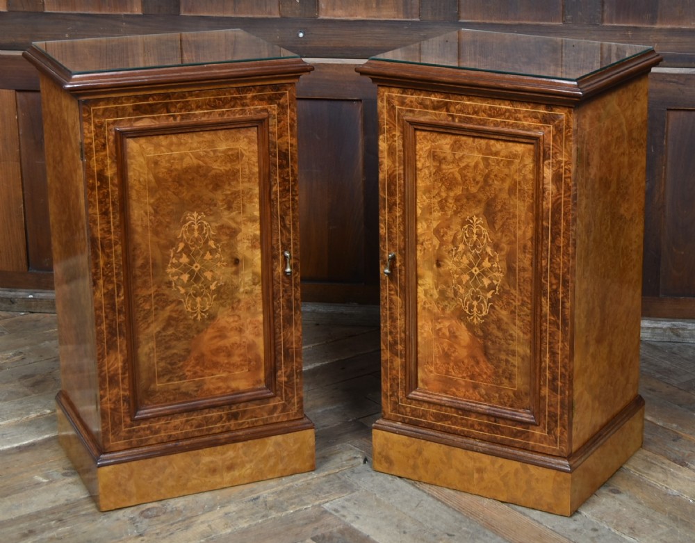 A matching pair of Edwardian burr walnut bedside pot cupboards featuring central marquetry inlays, single doors, glass tops, and plinth bases.