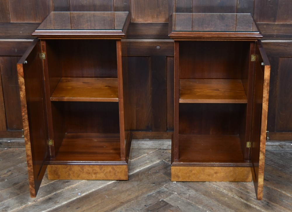 A matching pair of Edwardian burr walnut bedside pot cupboards featuring central marquetry inlays, single doors, glass tops, and plinth bases. View VI