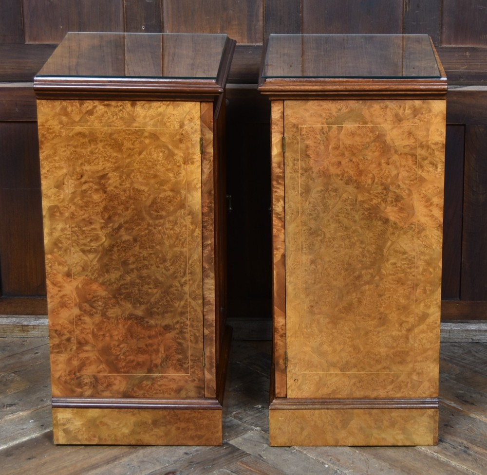 A matching pair of Edwardian burr walnut bedside pot cupboards featuring central marquetry inlays, single doors, glass tops, and plinth bases. View IX