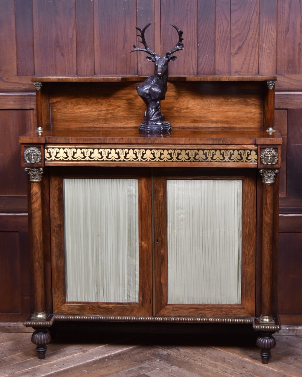 A Victorian dark rosewood chiffonier cabinet featuring a decorative brass inlay border, brass mask accents, two pleated silk fabric door panels, turned feet, and a raised upper tier shelf.