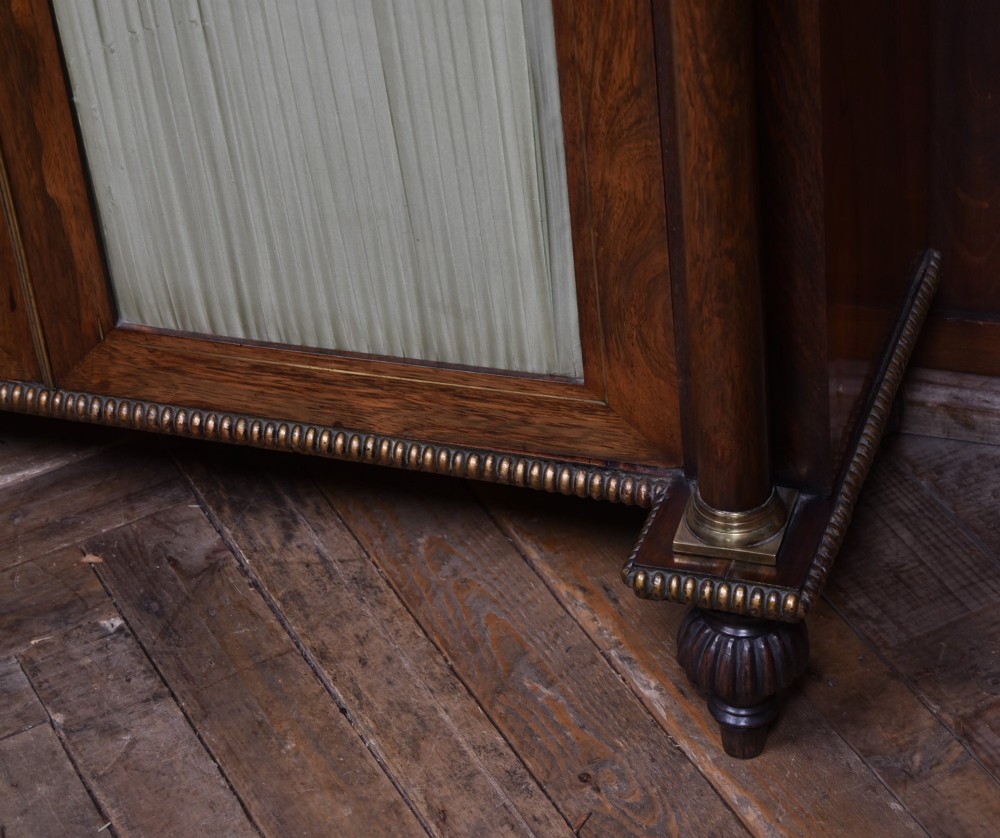 A Victorian dark rosewood chiffonier cabinet featuring a decorative brass inlay border, brass mask accents, two pleated silk fabric door panels, turned feet, and a raised upper tier shelf. View V