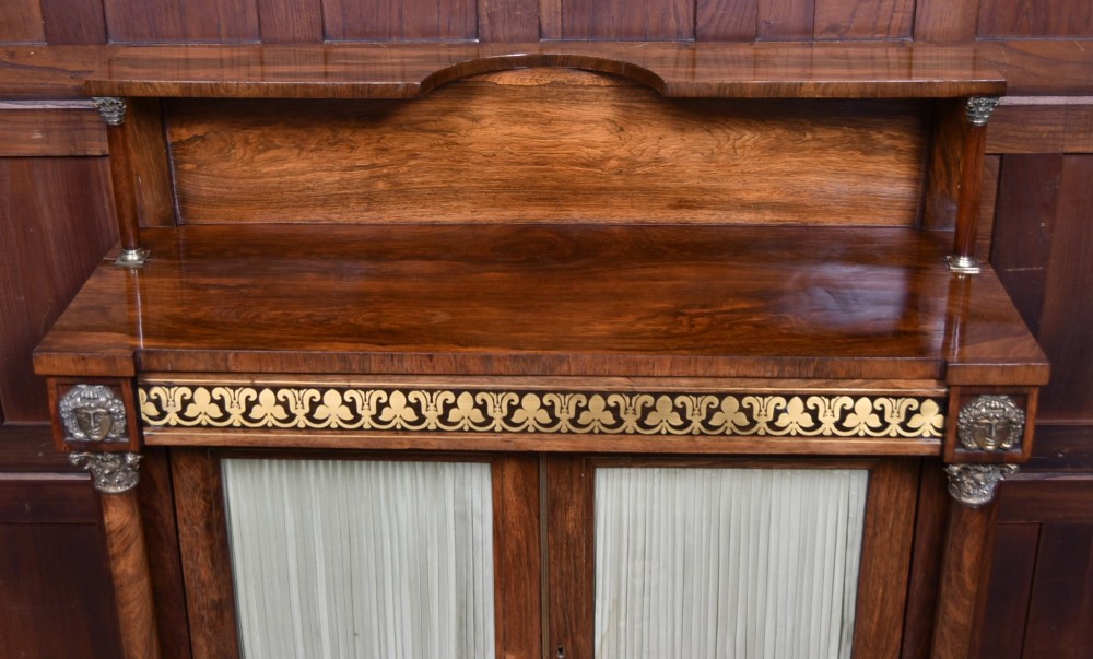 A Victorian dark rosewood chiffonier cabinet featuring a decorative brass inlay border, brass mask accents, two pleated silk fabric door panels, turned feet, and a raised upper tier shelf. View VI