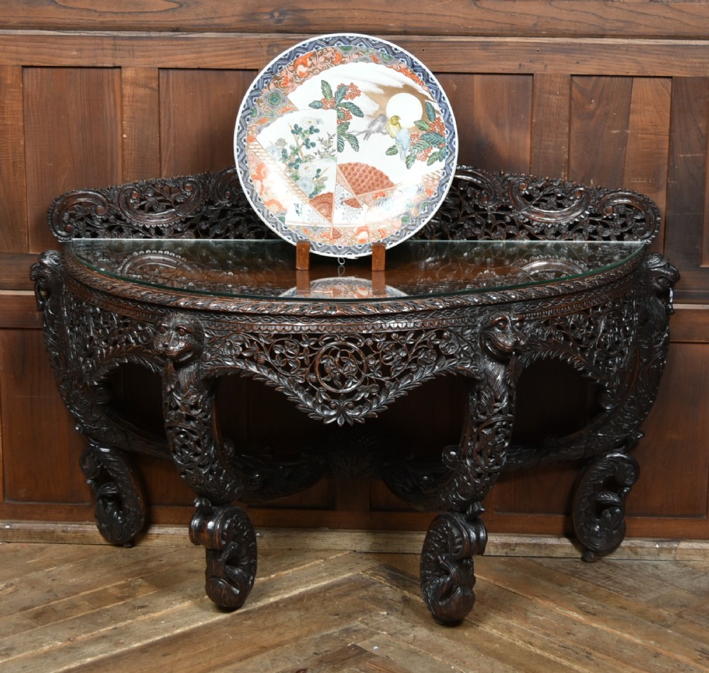 A dark hand-carved wooden demi-lune console table with intricate foliage openwork, carved lion mask legs, and a fitted glass top.