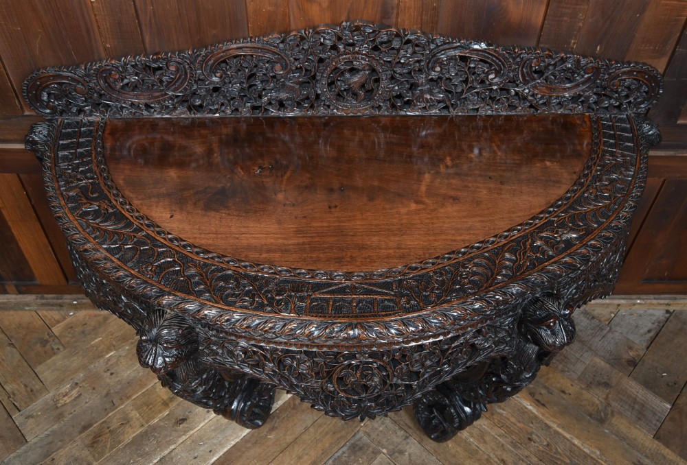 A dark hand-carved wooden demi-lune console table with intricate foliage openwork, carved lion mask legs, and a fitted glass top. View II