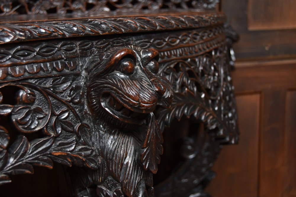 A dark hand-carved wooden demi-lune console table with intricate foliage openwork, carved lion mask legs, and a fitted glass top. View VIII