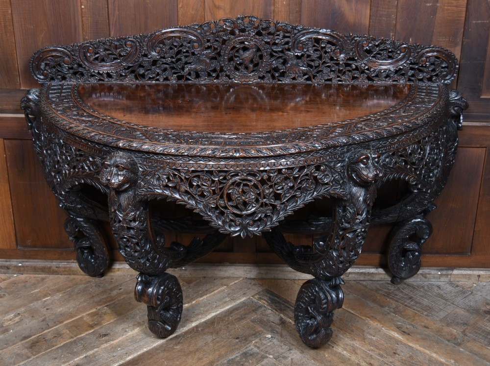 A dark hand-carved wooden demi-lune console table with intricate foliage openwork, carved lion mask legs, and a fitted glass top. View IX