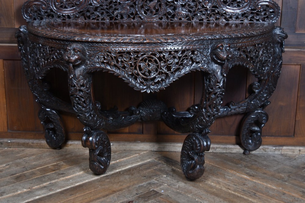 A dark hand-carved wooden demi-lune console table with intricate foliage openwork, carved lion mask legs, and a fitted glass top. View X