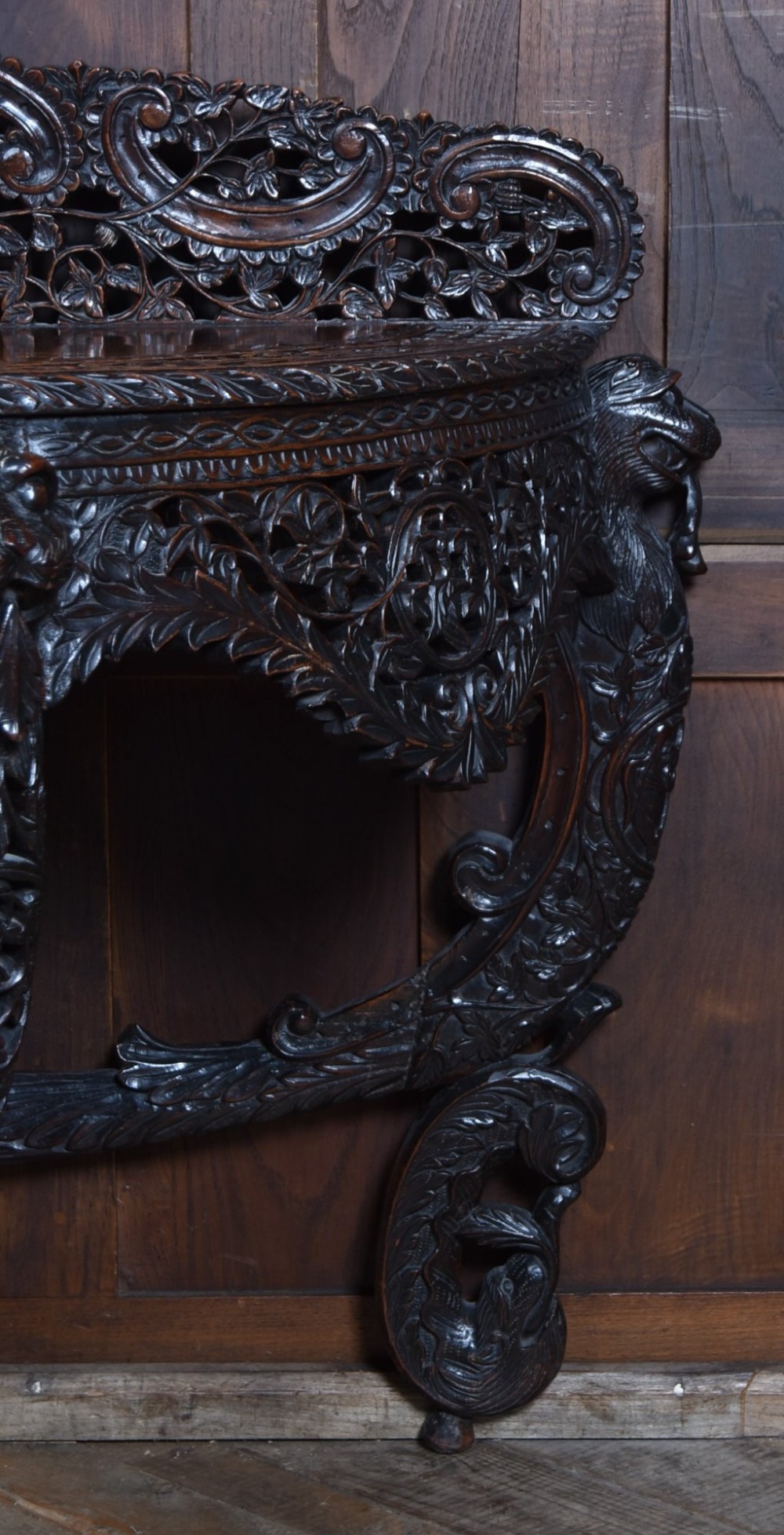 A dark hand-carved wooden demi-lune console table with intricate foliage openwork, carved lion mask legs, and a fitted glass top. View XI