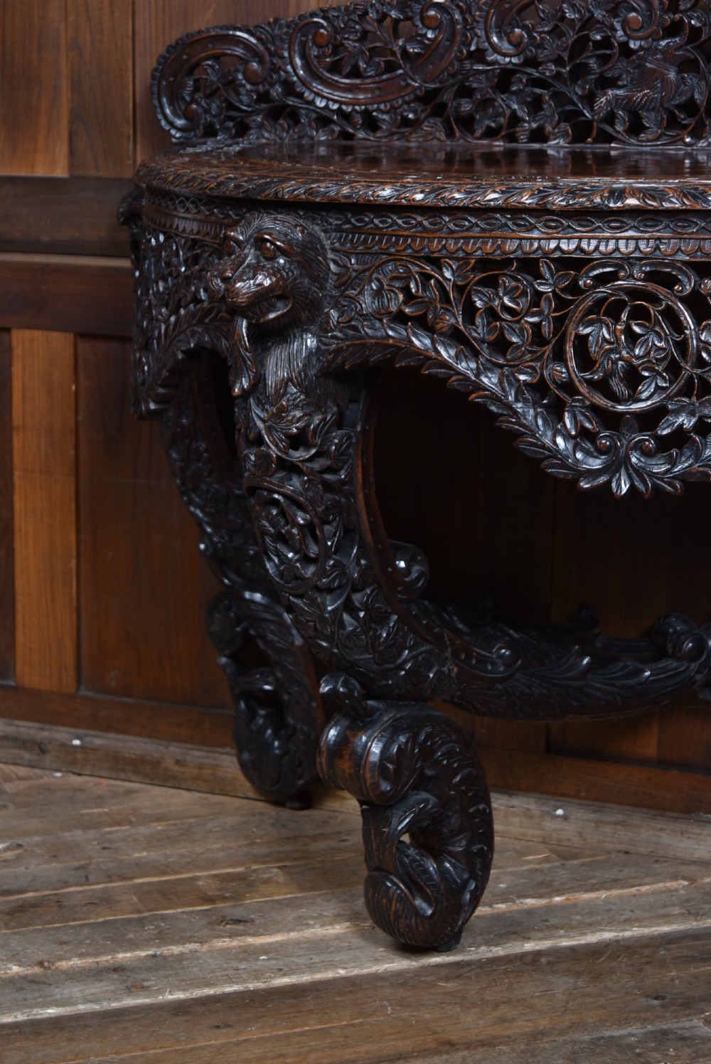 A dark hand-carved wooden demi-lune console table with intricate foliage openwork, carved lion mask legs, and a fitted glass top. View XII