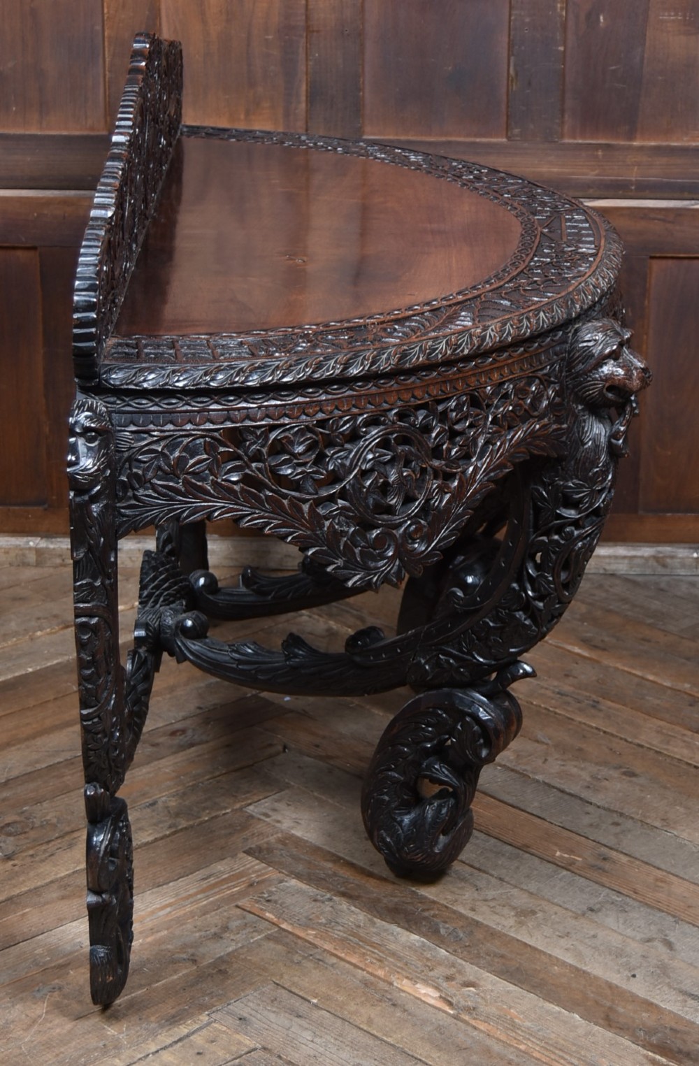 A dark hand-carved wooden demi-lune console table with intricate foliage openwork, carved lion mask legs, and a fitted glass top. View XIII