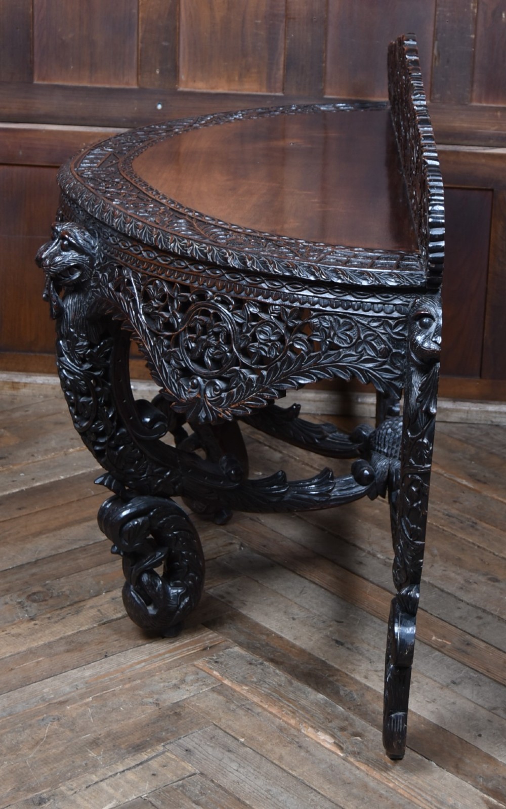 A dark hand-carved wooden demi-lune console table with intricate foliage openwork, carved lion mask legs, and a fitted glass top. View XV
