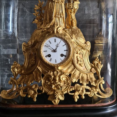 fig 2. An antique French gilt bronze mantel clock featuring a female figurative topper and Rococo scrollwork, housed inside a clear glass dome on a black oval base.