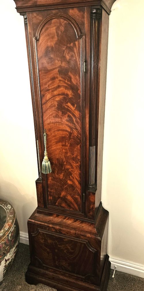 fig 3. A Georgian flame mahogany longcase clock featuring an arched hood with brass-capped fluted columns, an engraved brass dial with Roman numerals, and a trunk door showcasing rich wood grain.