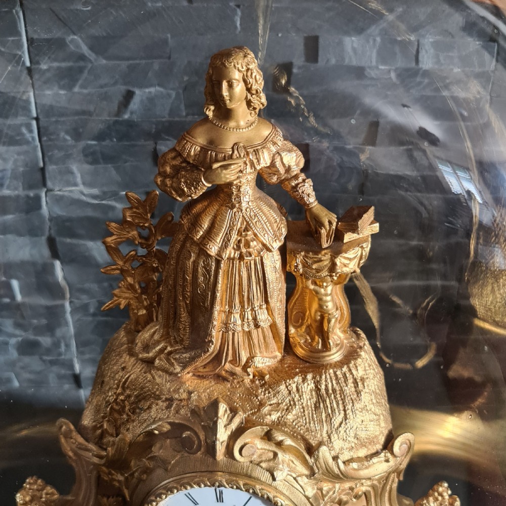 An antique French gilt bronze mantel clock featuring a female figurative topper and Rococo scrollwork, housed inside a clear glass dome on a black oval base. View II