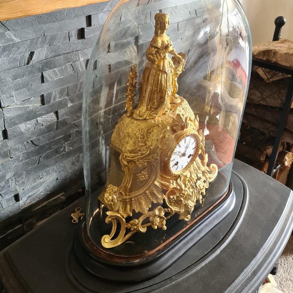 An antique French gilt bronze mantel clock featuring a female figurative topper and Rococo scrollwork, housed inside a clear glass dome on a black oval base. View IV