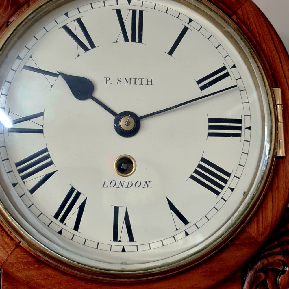 A petite mahogany drop dial wall clock featuring a cream dial with Roman numerals, framed by a brass bezel, carved side ears, and an inlaid floral motif on the lower trunk. View II