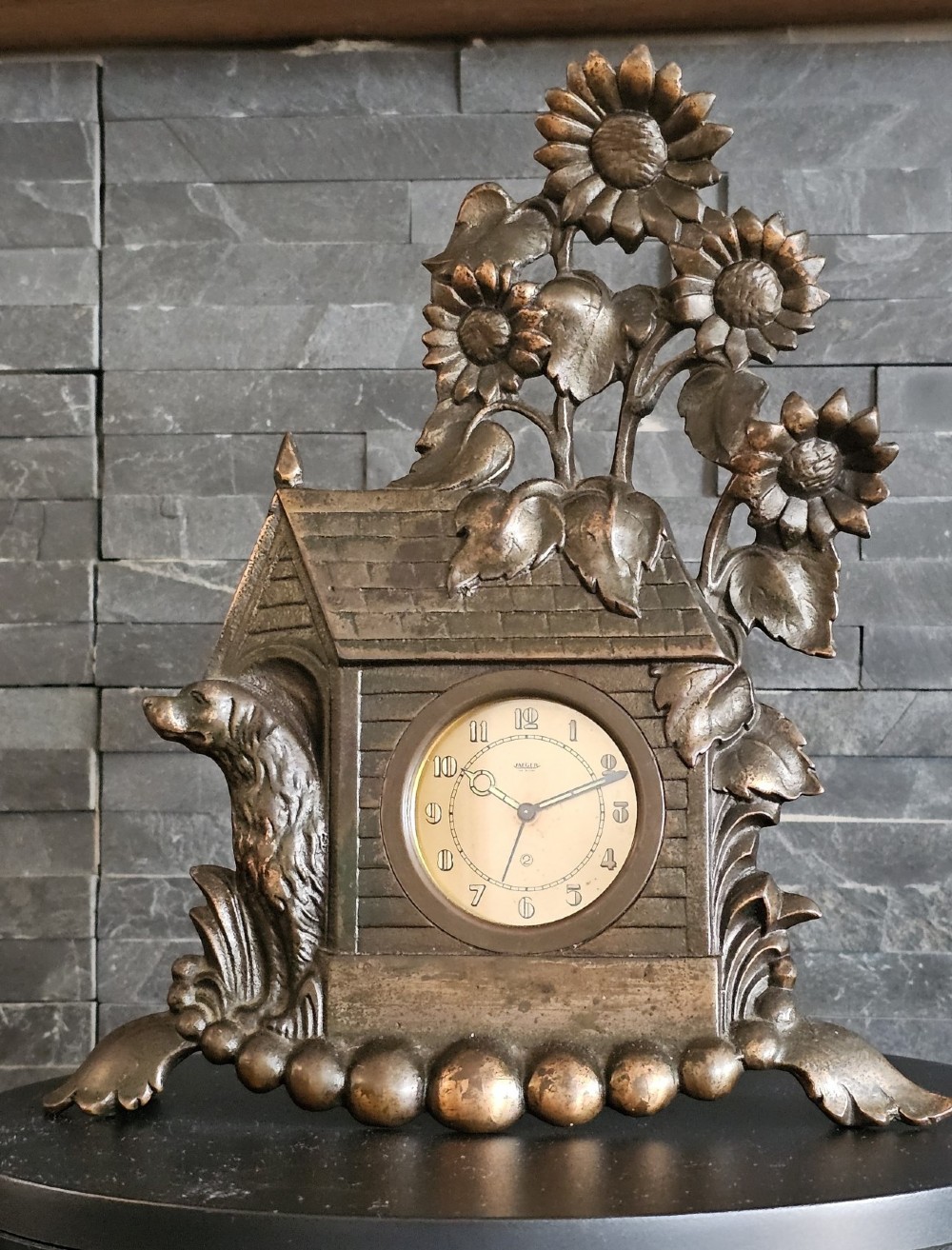 A bronzed Art Deco table clock shaped like a dog kennel with sunflowers on top and a dog figure on the left side of the circular dial.