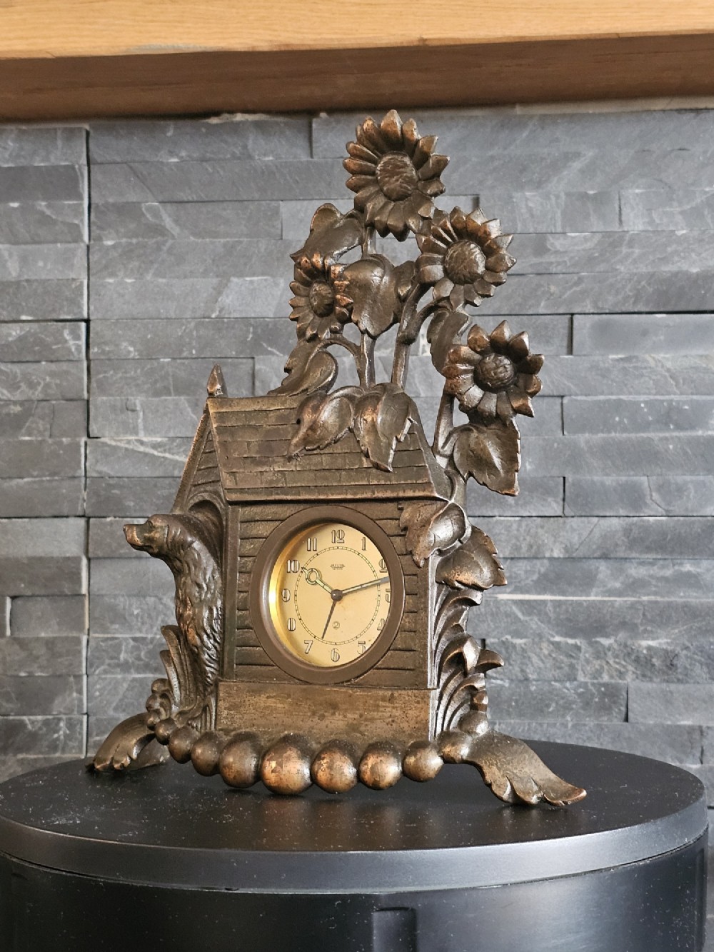 A bronzed Art Deco table clock shaped like a dog kennel with sunflowers on top and a dog figure on the left side of the circular dial. View II