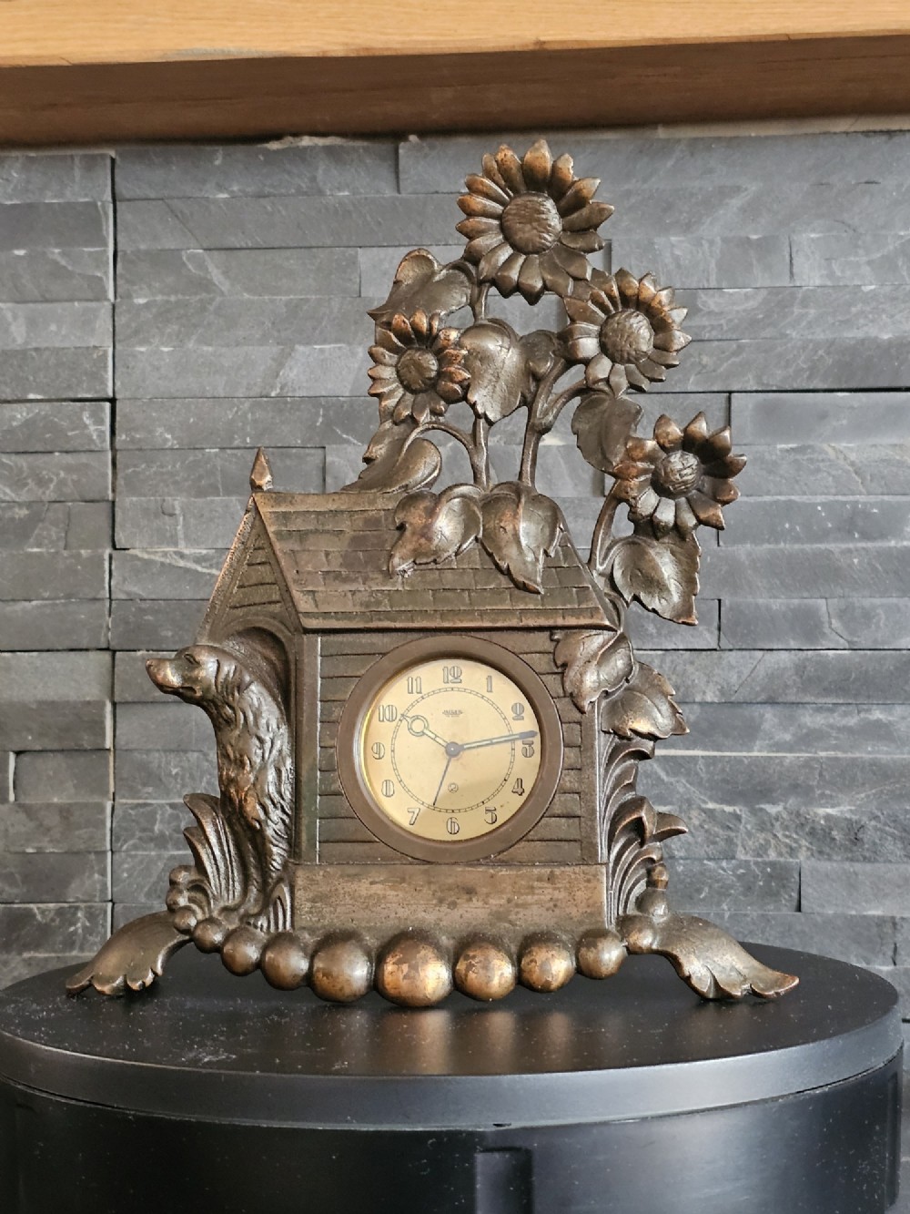 A bronzed Art Deco table clock shaped like a dog kennel with sunflowers on top and a dog figure on the left side of the circular dial. View V