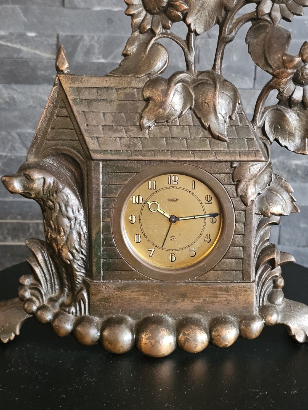 A bronzed Art Deco table clock shaped like a dog kennel with sunflowers on top and a dog figure on the left side of the circular dial. View IX