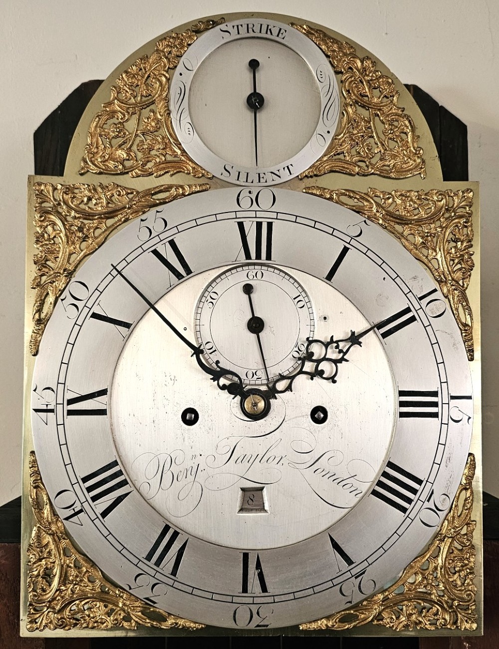 A Georgian flame mahogany longcase clock featuring an arched hood with brass-capped fluted columns, an engraved brass dial with Roman numerals, and a trunk door showcasing rich wood grain. View V