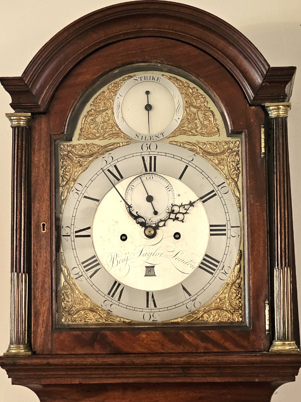 A Georgian flame mahogany longcase clock featuring an arched hood with brass-capped fluted columns, an engraved brass dial with Roman numerals, and a trunk door showcasing rich wood grain. View X