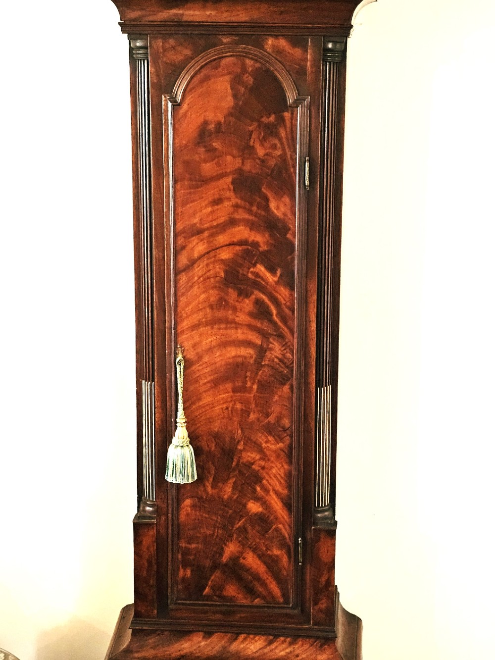 A Georgian flame mahogany longcase clock featuring an arched hood with brass-capped fluted columns, an engraved brass dial with Roman numerals, and a trunk door showcasing rich wood grain. View XI