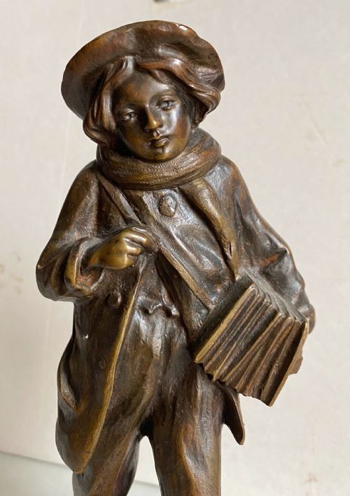 fig 1. A 19th-century patinated bronze sculpture depicting a schoolboy in a coat, cap, and scarf carrying a strap of books across his shoulder, standing on a cobblestone base.