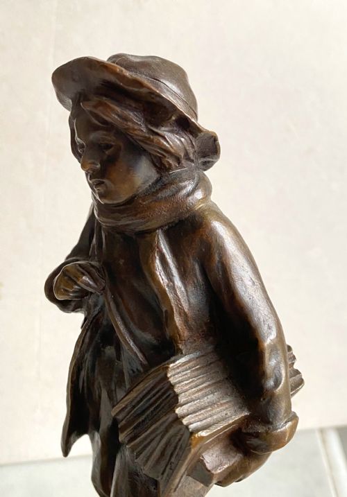 fig 2. A 19th-century patinated bronze sculpture depicting a schoolboy in a coat, cap, and scarf carrying a strap of books across his shoulder, standing on a cobblestone base.